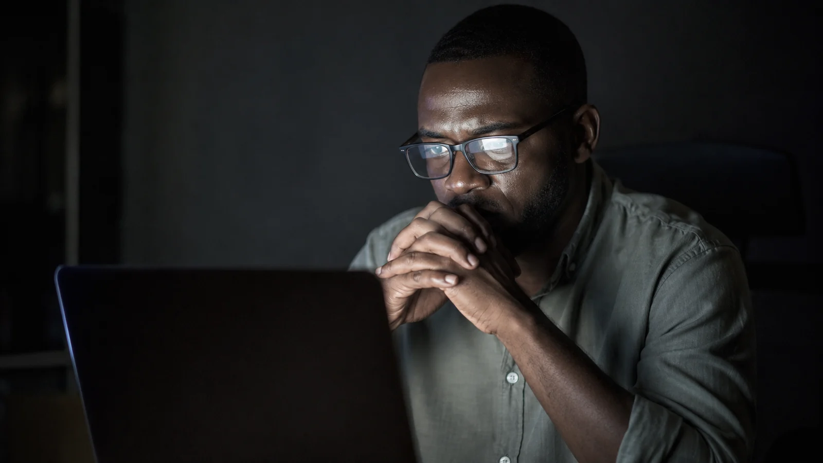 Dirigeant africain seul dans le noir, mains jointes devant la bouche, le reflet de son &eacute;cran d&rsquo;ordinateur dans ses lunettes